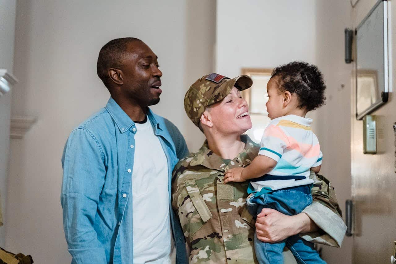 Smiling military woman in uniform holding child, with man beside them, celebrating family reunion, representing VA loan benefits for veterans and homebuyers.