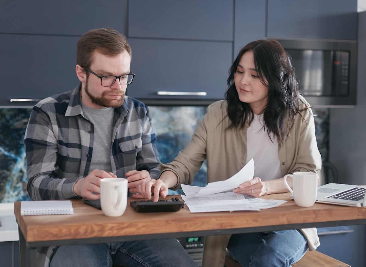 Couple discussing specialty mortgage loan options, reviewing documents and using a calculator at a kitchen table, emphasizing financial planning and guidance.